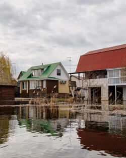 Several homes, are partially submerged in floodwater, indicating significant flooding in the area. The sky is overcast, and the surroundings show signs of flood damage, with debris and plants in the water.