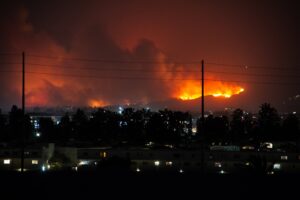 Distant view of a wildfire at night, with bright flames and smoke illuminating the sky. Below, a cityscape with lit buildings is visible in the foreground.
